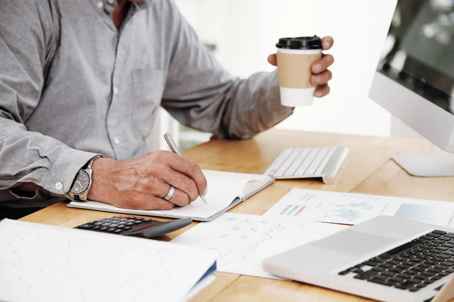 An advisor writing notes at a desk beside a laptop, calculator, and printed financial charts.